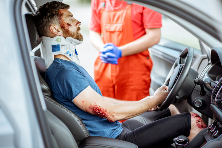 Un hombre está herido con graves heridas y un collarin en el cuello sentado en el asiento del conductor esperando como le atienden los médicos.